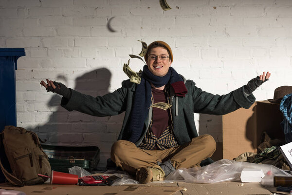 happy homeless man tossing up money while sitting on rubbish dump