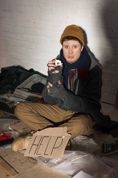 угнетенный бездомный сидит рядом с картонной карточкой с рукописным текстом "Помогите" и держит дом для покроя бумаги

