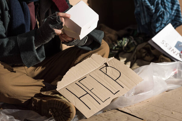 partial view oh homeless man in fingerless gloves sitting near cardboard card with "help" inscription and holding paper box