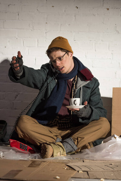 Бездомный держит чашку кофе и пользуется винтажным телефоном
