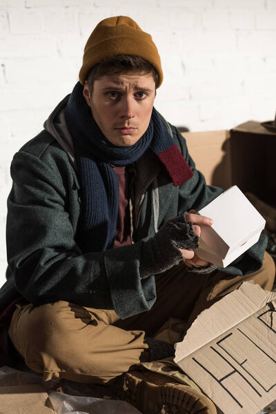 sad homeless man sitting with cardboard card with "help" lettering and holding white paper box