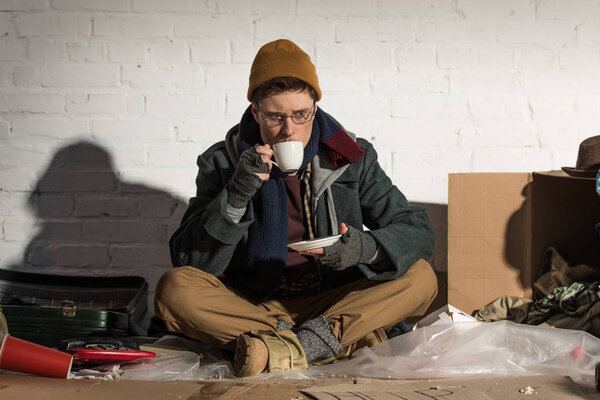 Бездомный пьет кофе, сидя у стены из белого кирпича на свалке.
