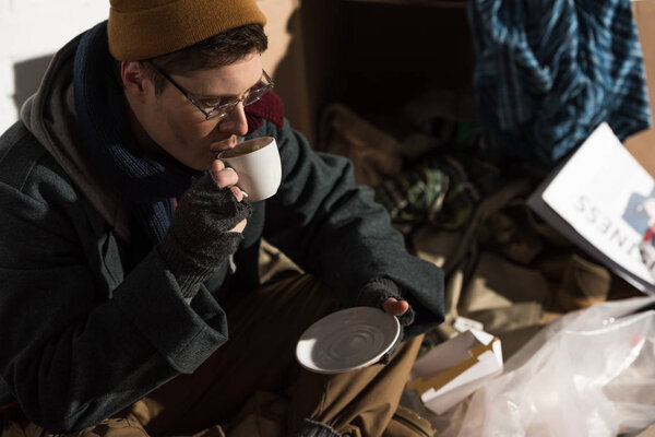 Бездомный в очках и перчатках без пальцев пьет кофе
