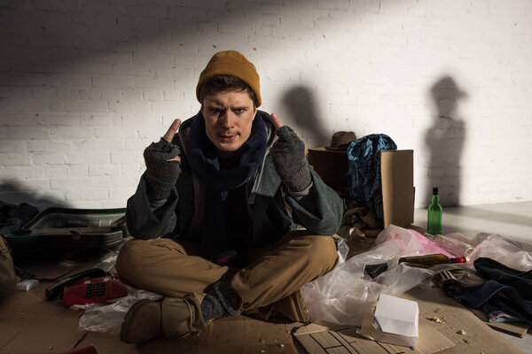 homeless man showing middle fingers while sitting surrounded by rubbish