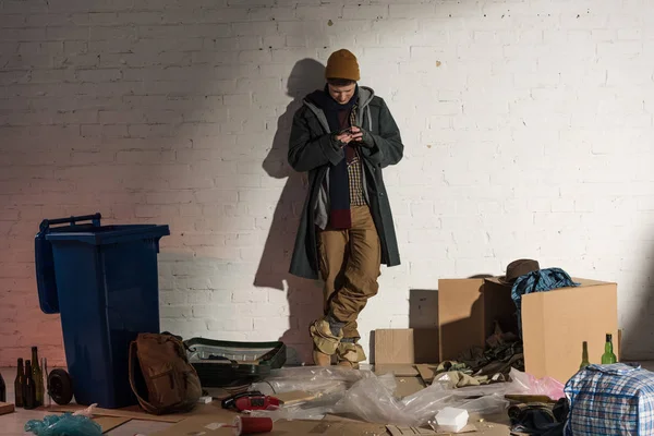 Άστεγος Χρησιμοποιώντας Smartphone Ενώ Στέκεται Λευκό Τοίχο Περιβάλλεται Από Σκουπίδια — Φωτογραφία Αρχείου