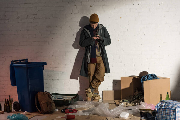 homeless man using smartphone while standing by white brick wall surrounded by rubbish