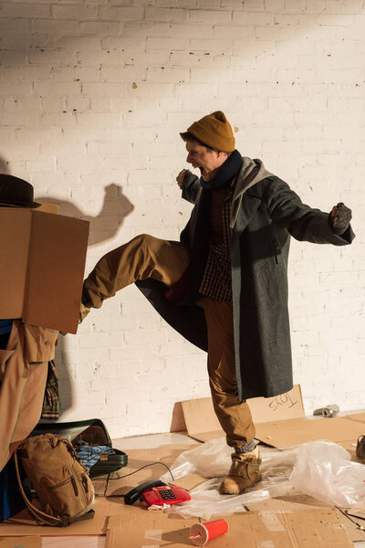 aggressive screaming homeless man kicking trash container with cardboard box
