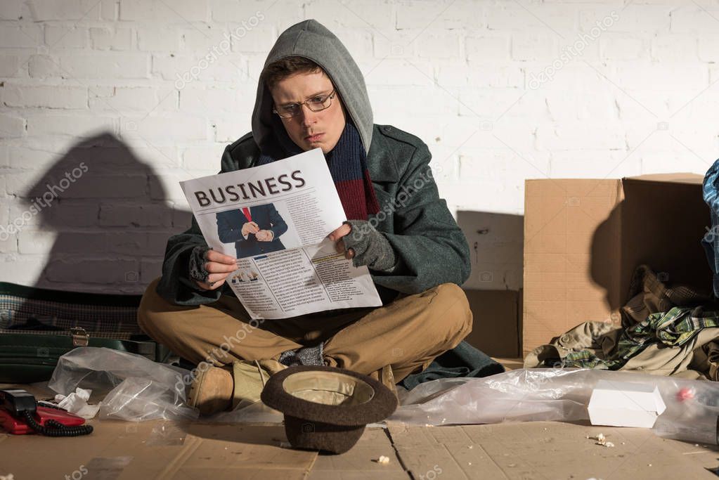 homeless beggar reading business newspaper while sitting by brick wall