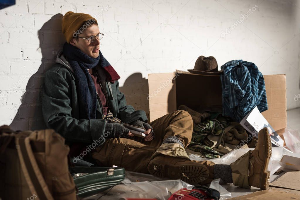 homeless man sitting by brick wall surrounded by rubbish