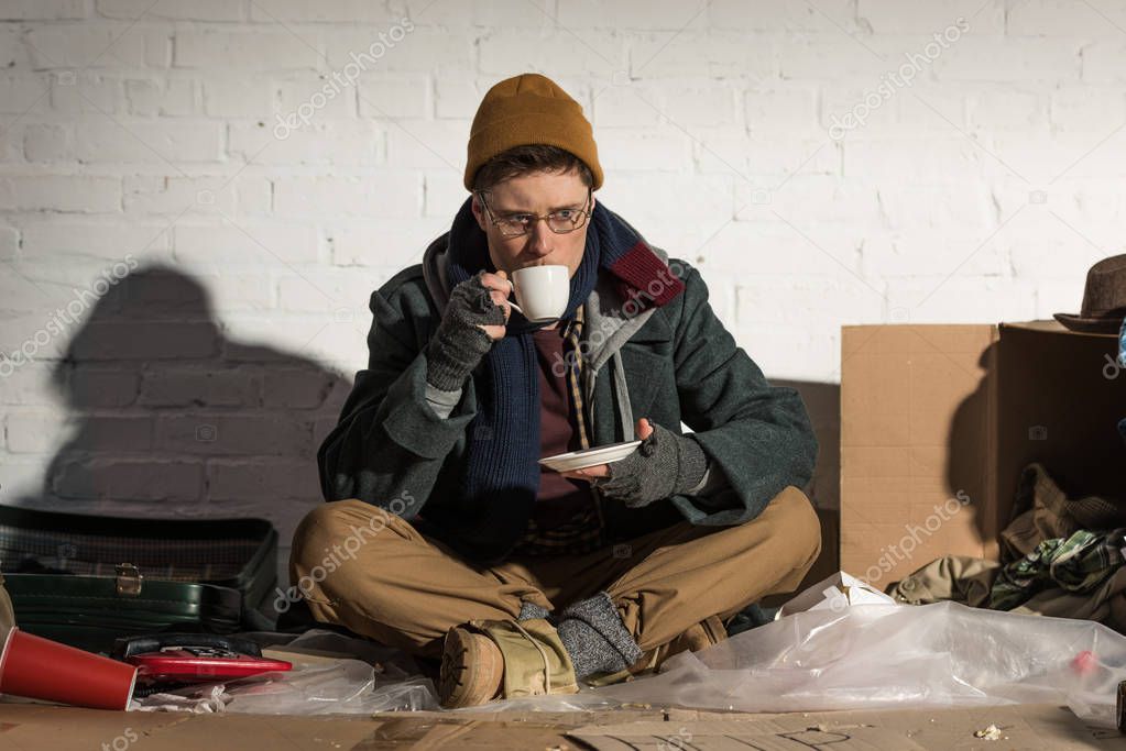 homeless man drinking coffee while sitting by white brick wall on rubbish dump