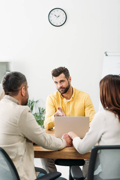 selective focus of advisor sitting at table and pointing with hand at laptop near man and woman in office