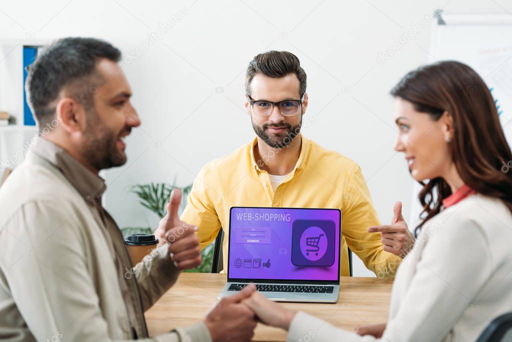 selective focus of advisor pointing with finger at laptop with web shopping website on screen while investor holding woman hand and thumbing up in office