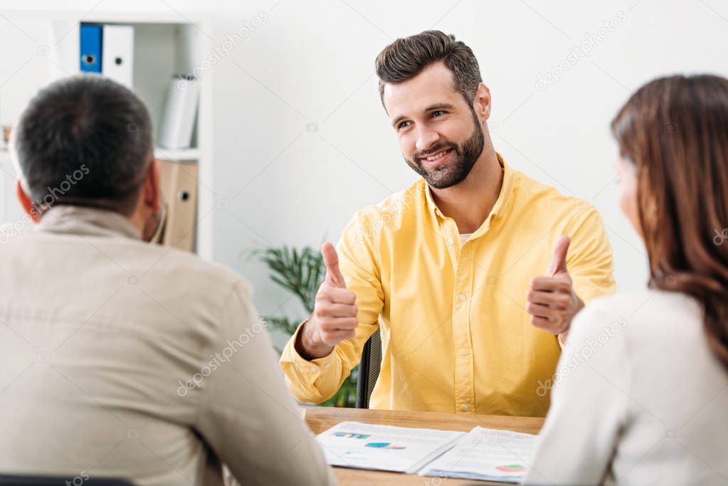 selective focus of advisor sitting at table and thumbing ups near man and woman in office