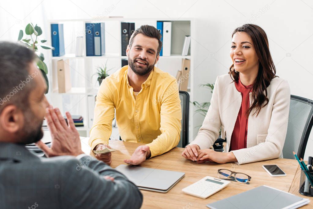 selective focus of investor sitting at table near woman and giving dollar banknote to advisor in office