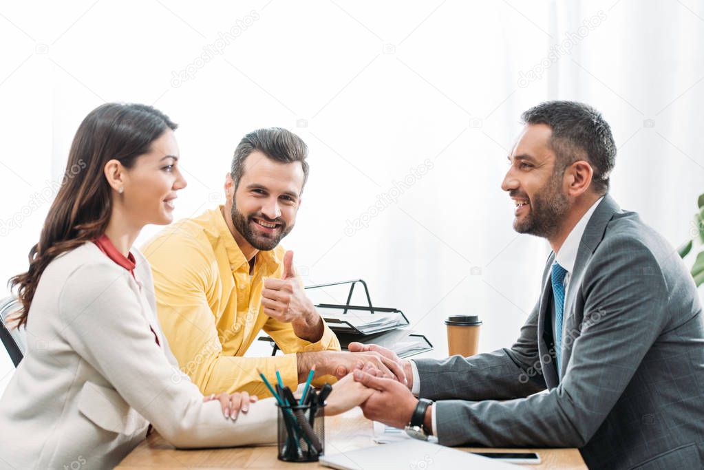 advisor sitting at table and holding investors hands wile man thumbing up in office