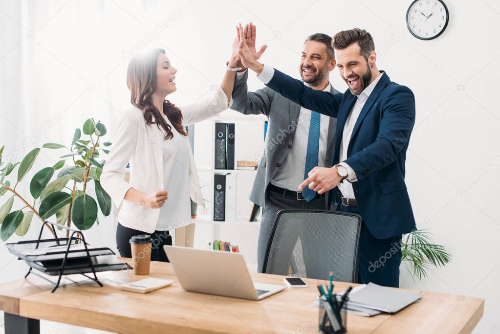 colleagues standing near table with laptop and highing five in office