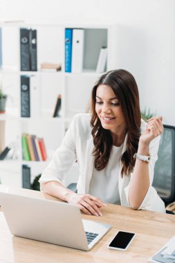 masada oturan, laptop için arıyorum ve ofiste gülümseyen iş kadını