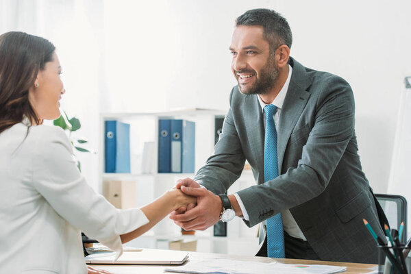 handsome advisor in suit shaking hands with investor at workplace  