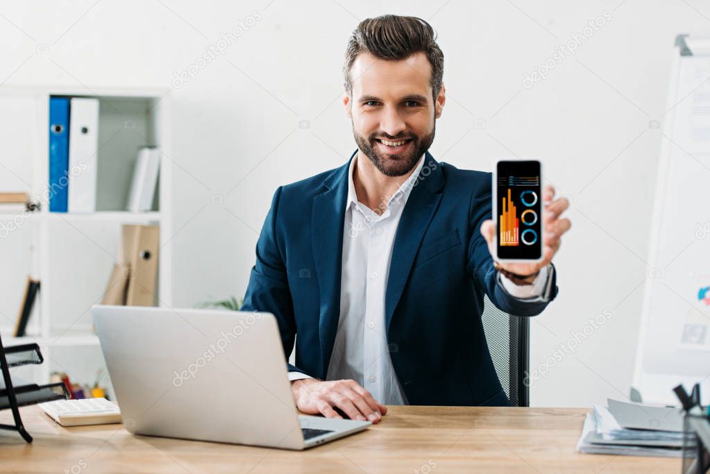 businessman sitting at table with laptop and showing smartphone with charts and graphs app on screen in office