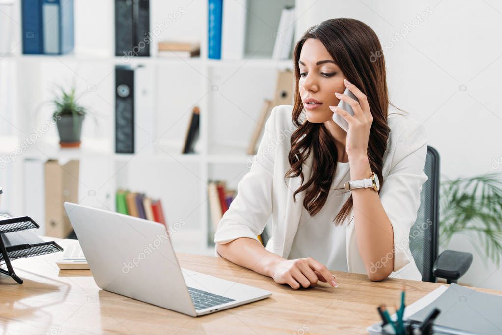 businesswoman sitting at table, looking to laptop and talking on smartphone in office