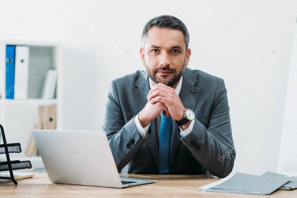 handsome businessman sitting at table with laptop and looking at camera in office