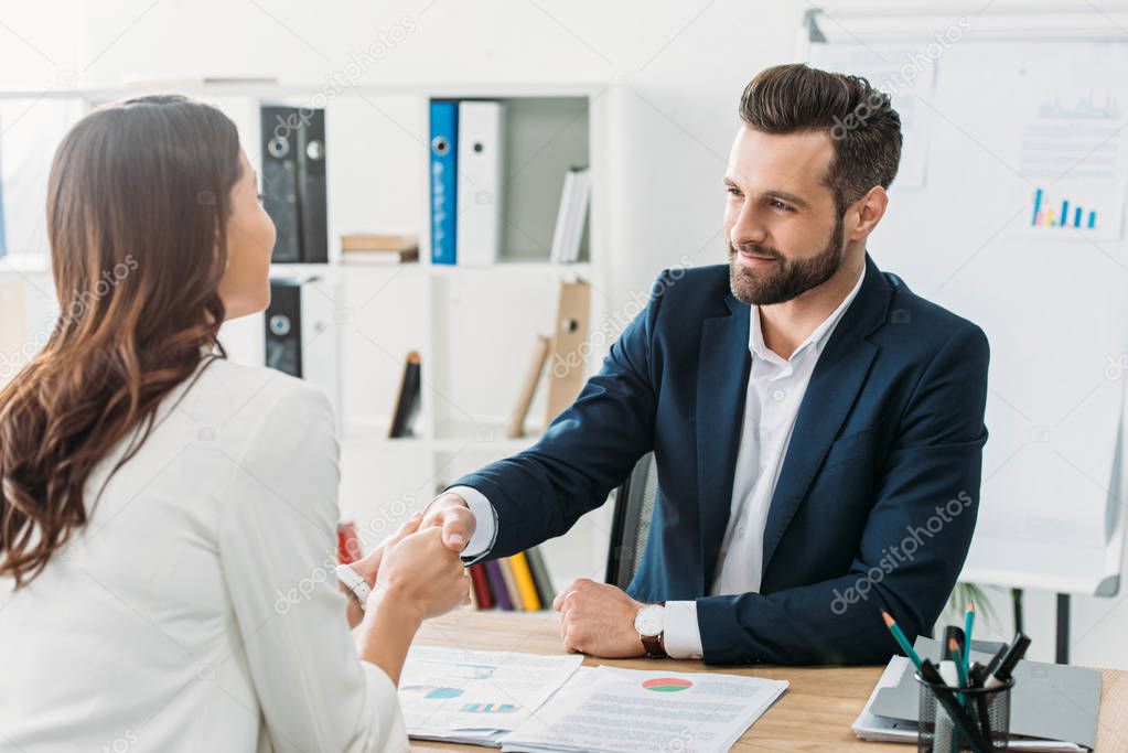 handsome advisor in suit shaking hands with investor at workspace