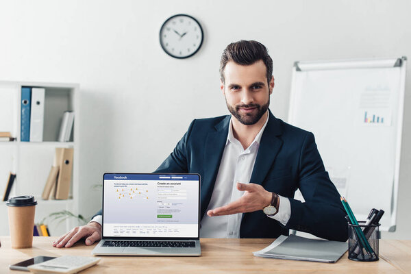handsome advisor in suit pointing with fingers at laptop with facebook website on screen at office 