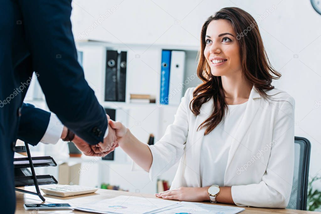 beautiful and smiling advisor and investor in suits shaking hands at workplace 