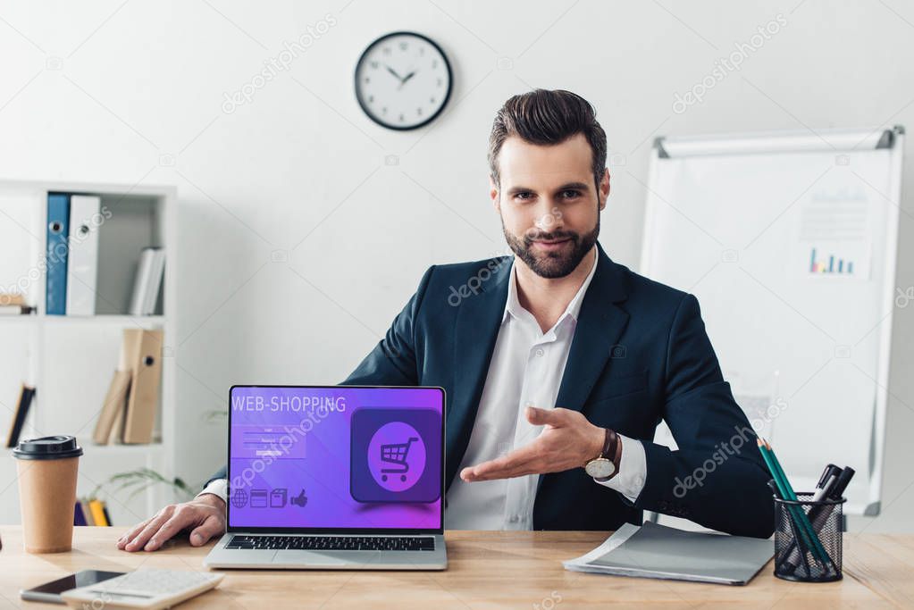 handsome advisor in suit pointing with fingers at laptop with web-shopping website on screen 