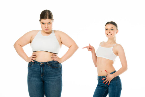 smiling fit woman pointing with finger at overweight angry woman isolated on white