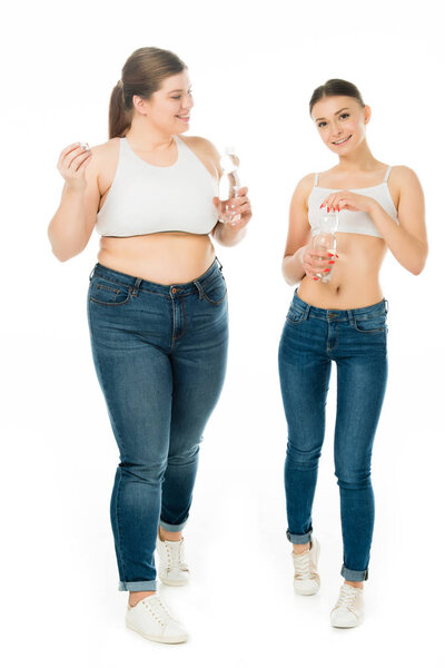 smiling slim and overweight women in jeans holding bottles with water isolated on white