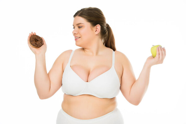 happy young overweight woman holding doughnut and green apple isolated on white