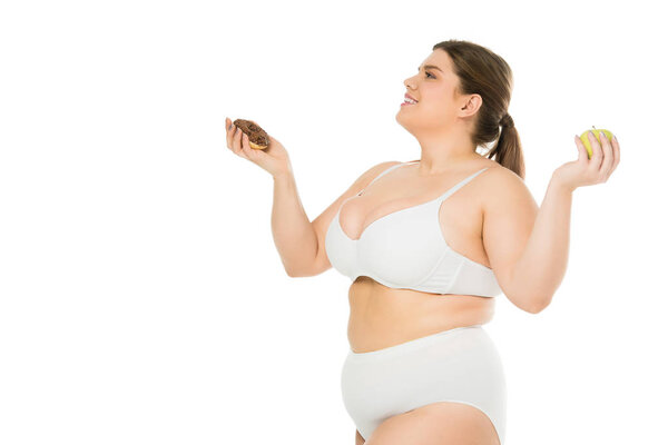 smiling young overweight woman holding doughnut and green apple isolated on white
