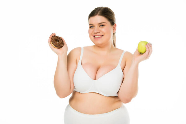 cheerful young overweight woman holding doughnut and green apple isolated on white