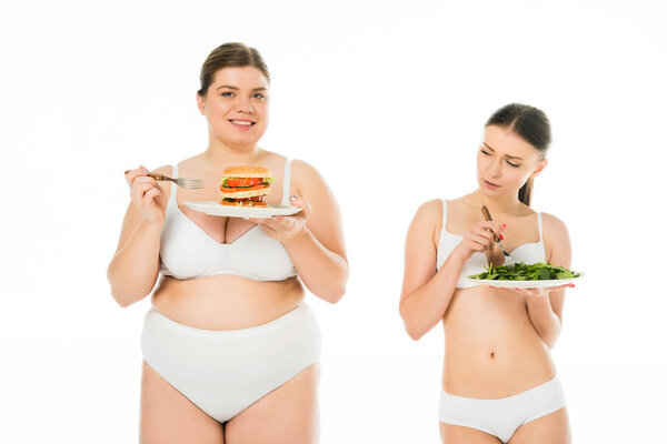 upset slim woman standing with plate of green spinach leaves while smiling overweight woman holding plate with burger isolated on white