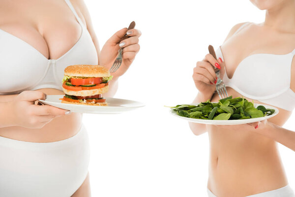 cropped view of slim woman standing with plate of green spinach leaves while overweight woman holding plate with burger isolated on white