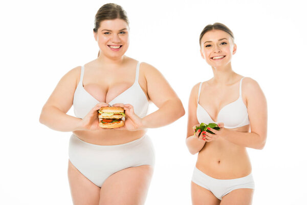 slim happy woman in underwear holding green spinach leaves while overweight smiling woman holding burger isolated on white