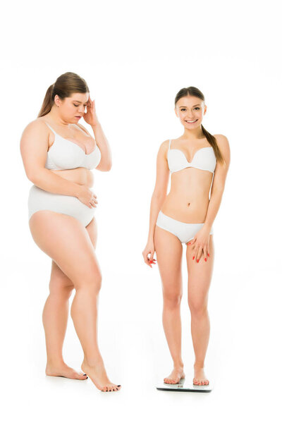 shocked overweight woman in underwear looking at happy slim woman on scales isolated on white, body positivity concept