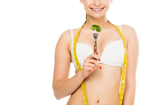 cropped view of happy slim woman with measuring tape holding fork with green spinach leaves isolated on white, dieting concept