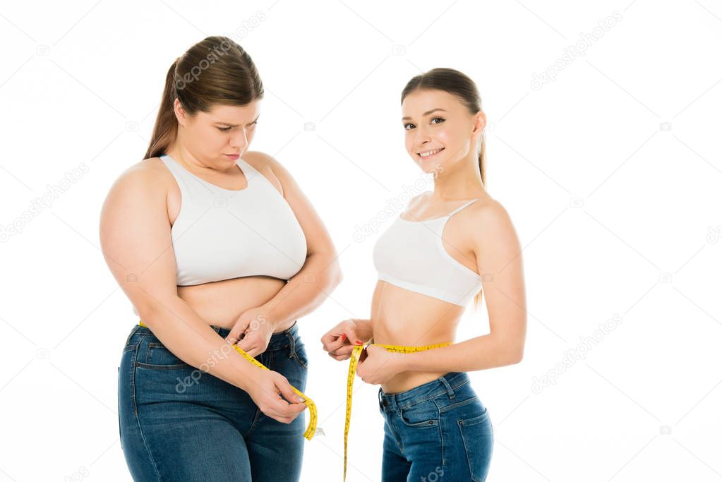 jealous overweight woman with measuring tape looking at slim smiling attractive woman isolated on white