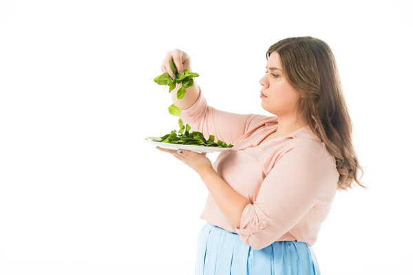 side view of overweight woman holding fresh green spinach leaves above plate isolated on white