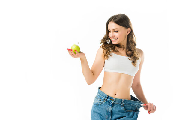 happy slim young woman in big jeans holding green apple isolated on white