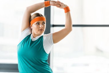 overweight woman stretching while standing in sportswear in gym clipart