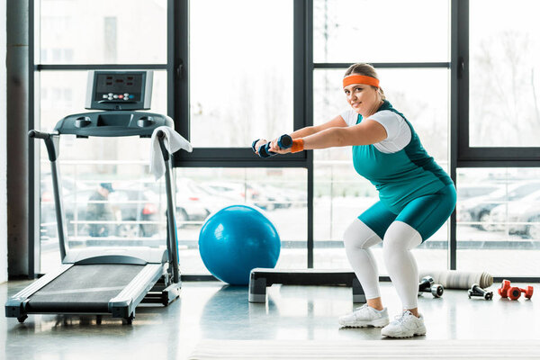 happy plus size woman exercising with dumbbells near treadmill 