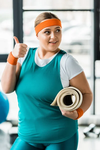 愉快的超重的女人显示拇指 而举行健身垫在健身房 — 图库照片
