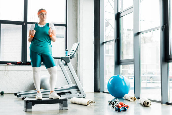 plus size woman standing on step platform near dumbbells in gym