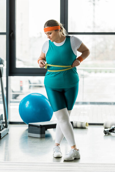 overweight girl looking at measuring tape while measuring waist in gym