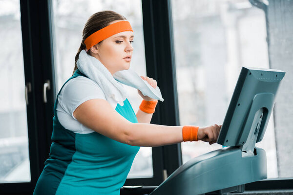 уставшая толстая девушка с полотенцем на плечах тренируется на беговой дорожке в тренажерном зале

