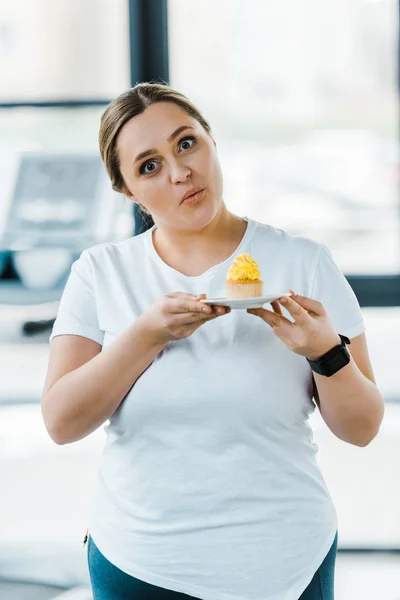惊讶超重的女人拿着美味的纸杯蛋糕在健身房 — 图库照片