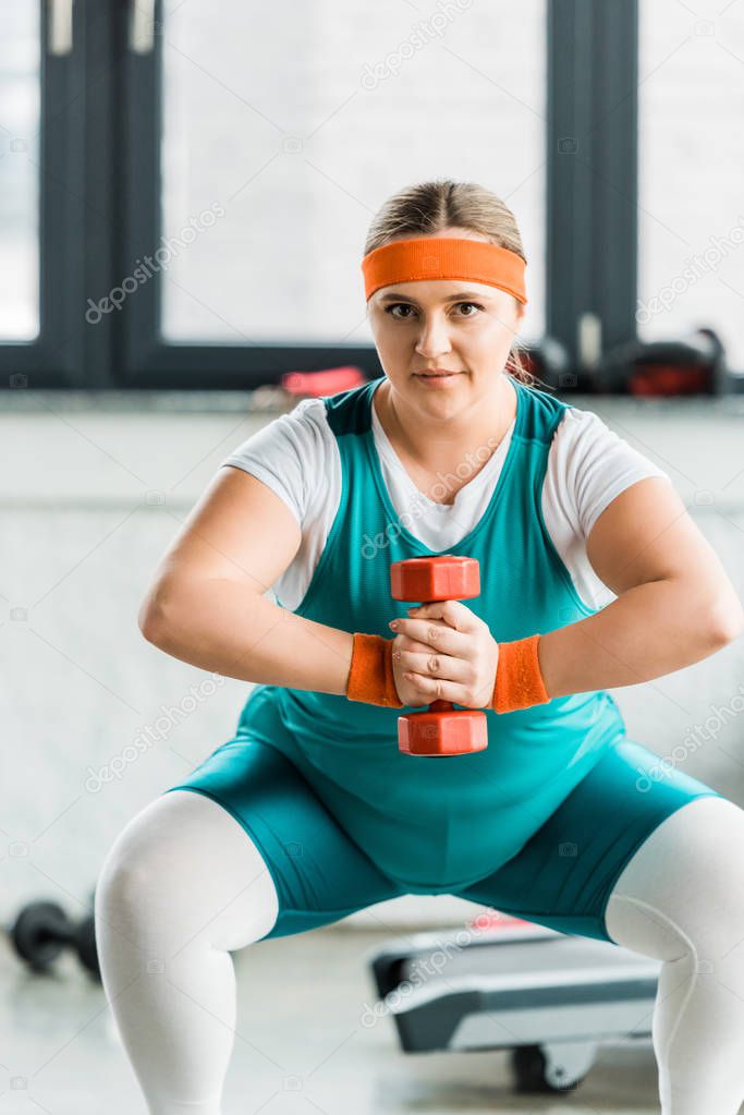 attractive overweight girl in sportswear squatting with dumbbell in gym